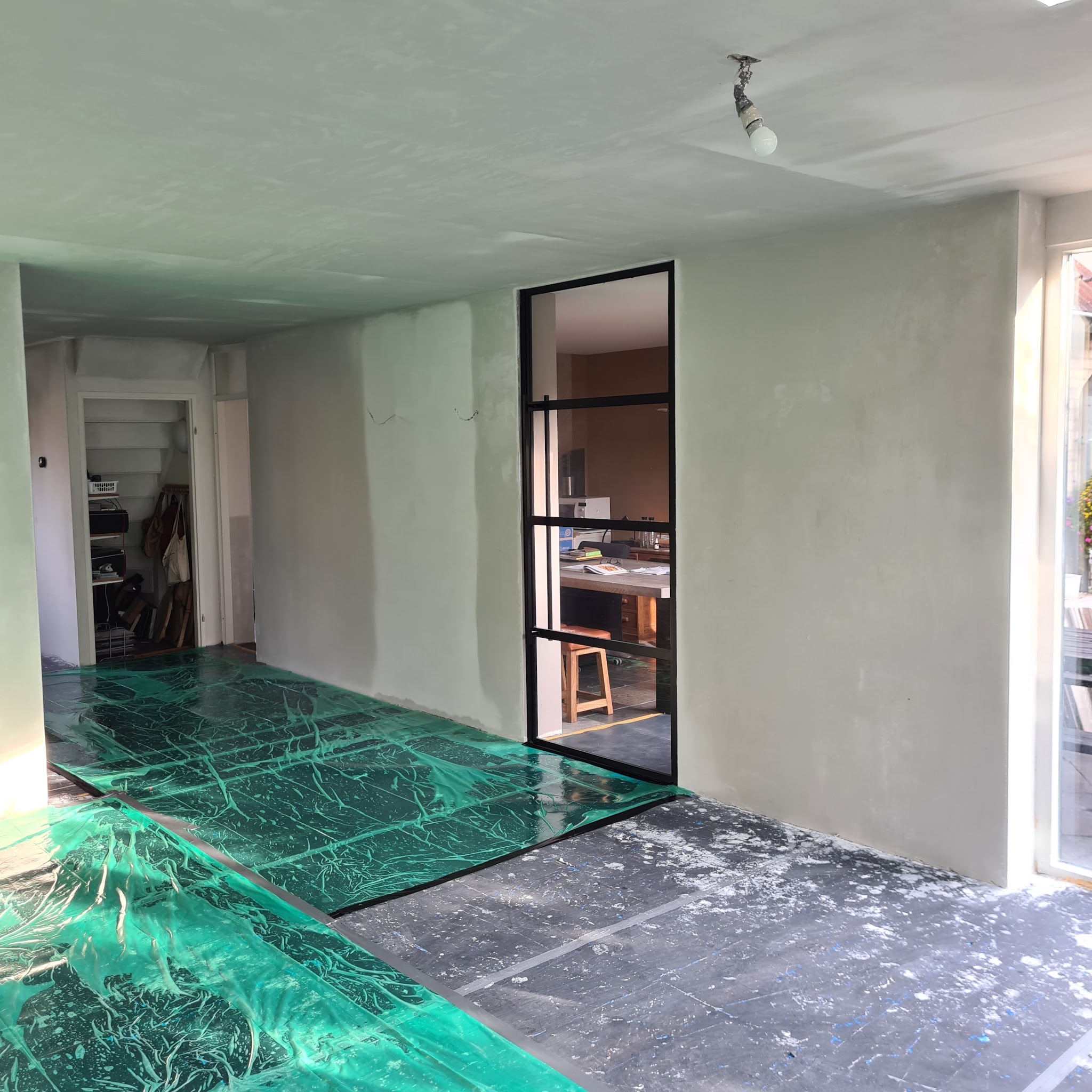 Vers gestuukte wanden en plafond in een woonkamer met stalen deur, vloer afgedekt met groen folie